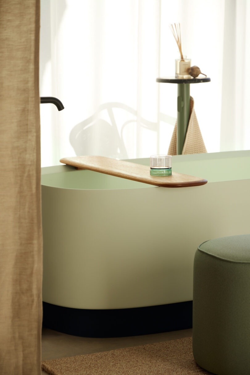 Minimalist bathroom with green bathtub, wooden tray holding a small candle, green ottoman, hanging towel, and thin table near a sheer curtain