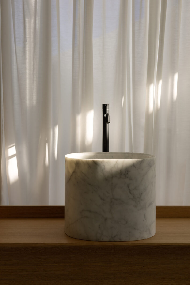 Marble sink with a black faucet on a wooden counter, set against sheer white curtains