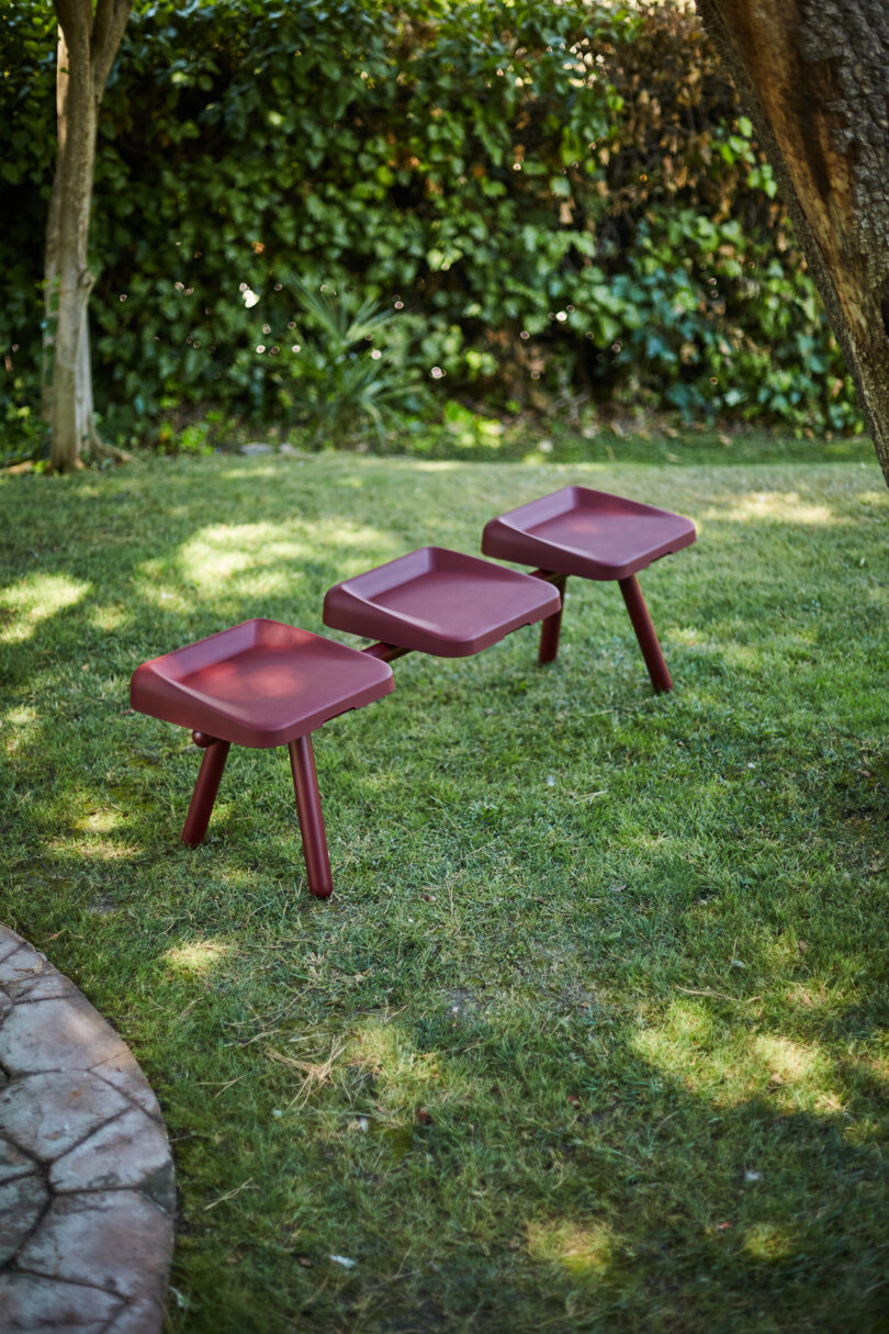 Three red, square stools with red legs are arranged in a row on a grassy lawn with trees and greenery in the background