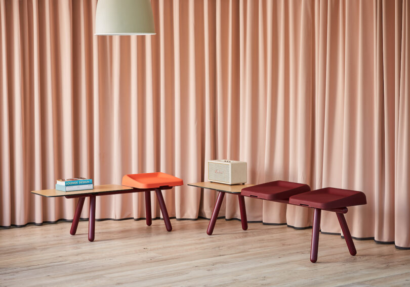 Two modern benches with unique, colorful cushions are placed in front of beige curtains. A lamp hangs from the ceiling above, illuminating the scene