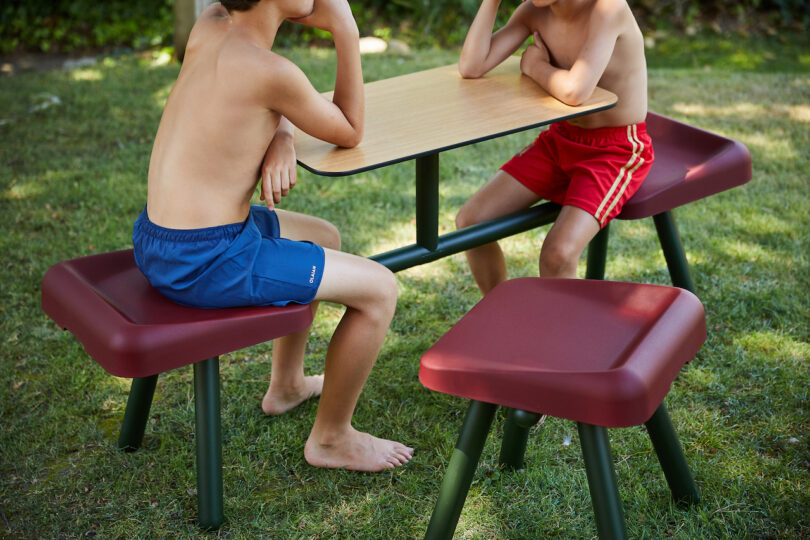 Two shirtless individuals in blue and red shorts sit at an outdoor table on the grass