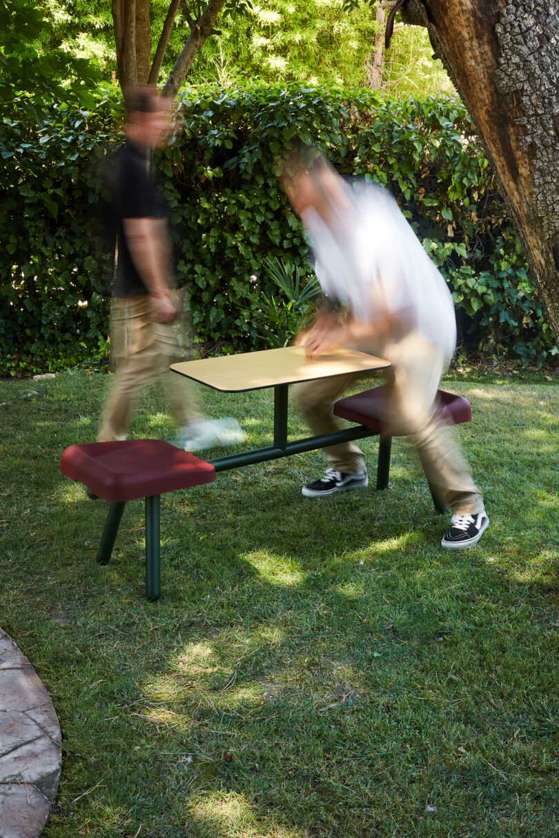 Two people in motion near a picnic table with attached benches, in a grassy outdoor setting surrounded by trees and shrubs