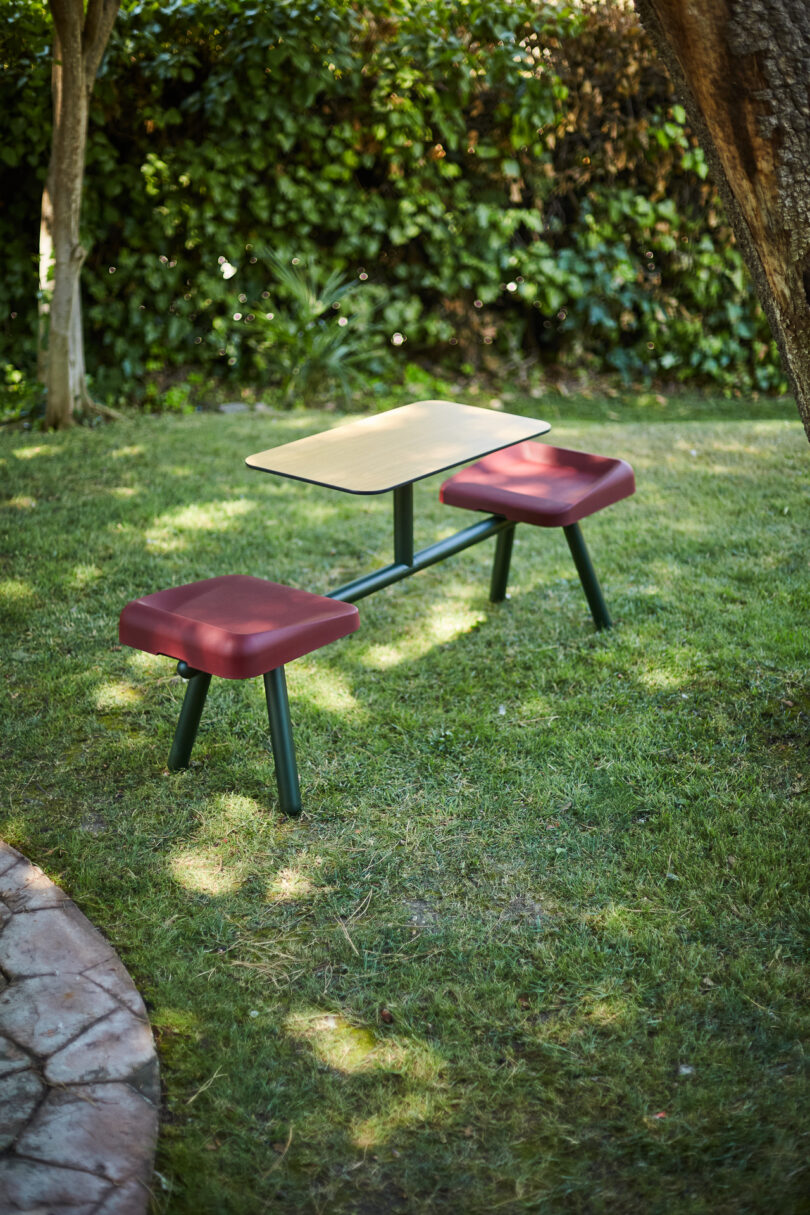 A small outdoor table with two attached red cushioned seats on a grassy area, surrounded by trees and shrubbery