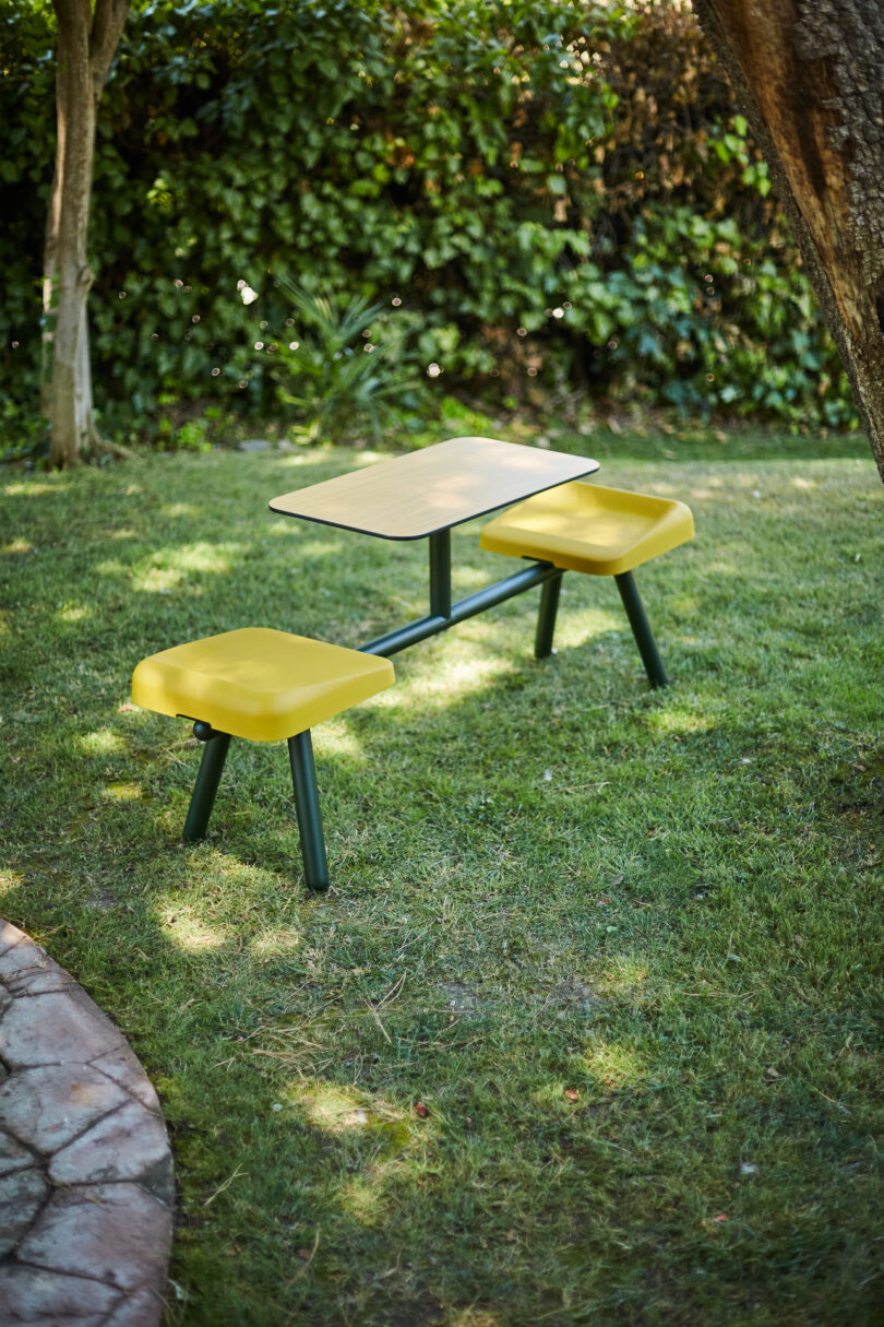 A small outdoor table with two attached yellow cushioned seats on a grassy area, surrounded by trees and shrubbery