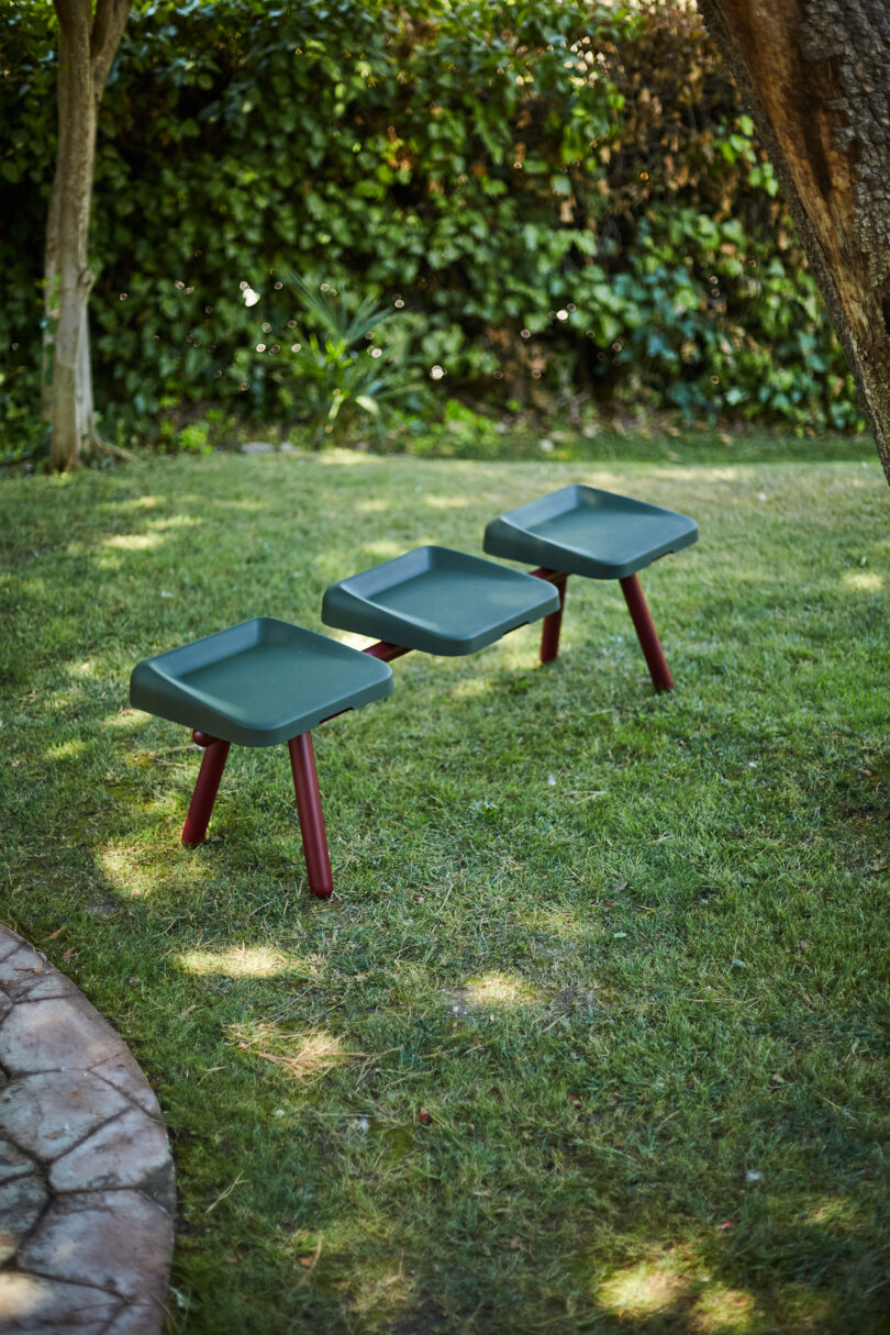 Three green, square stools with red legs are arranged in a row on a grassy lawn with trees and greenery in the background