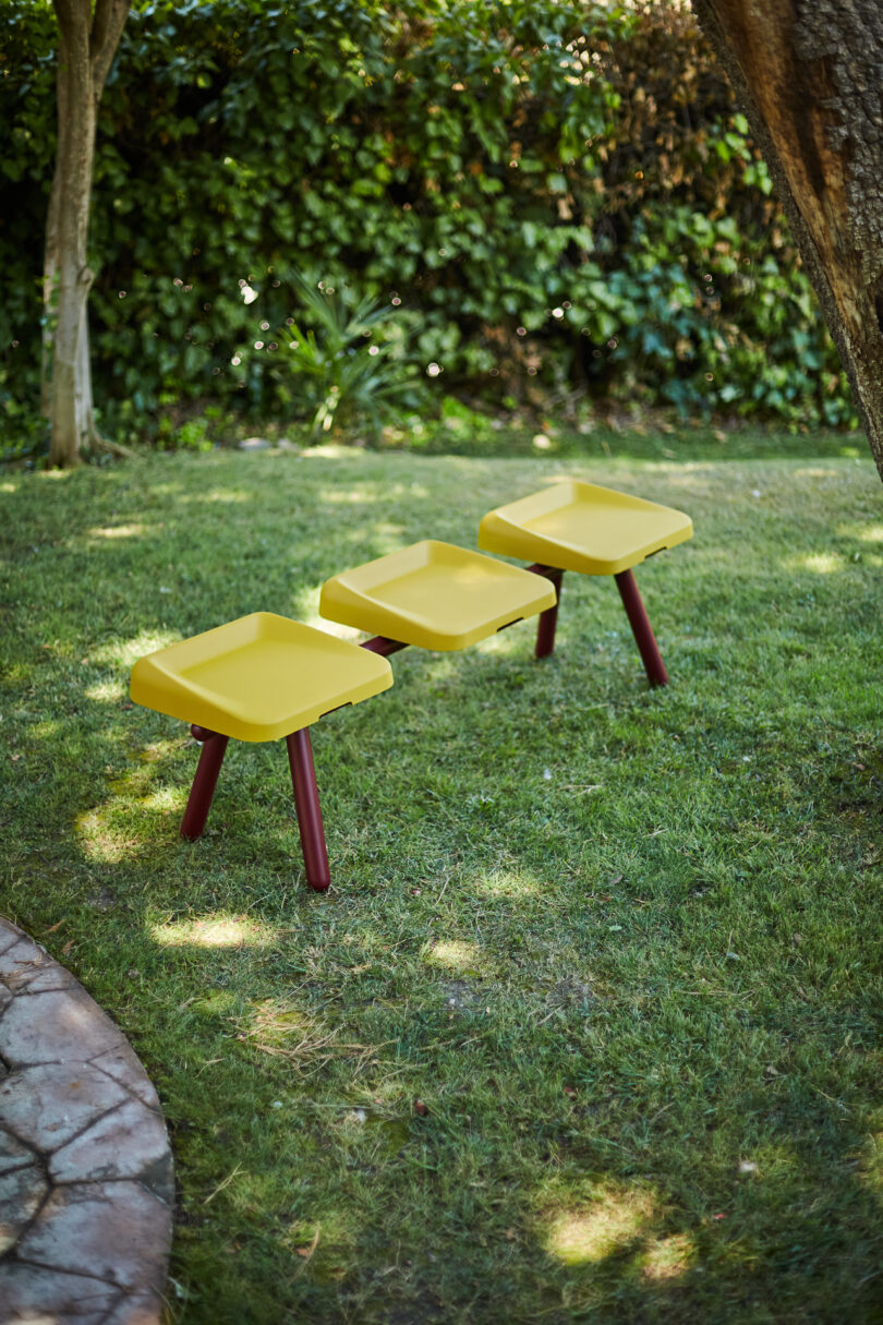 Three yellow, square stools with red legs are arranged in a row on a grassy lawn with trees and greenery in the background