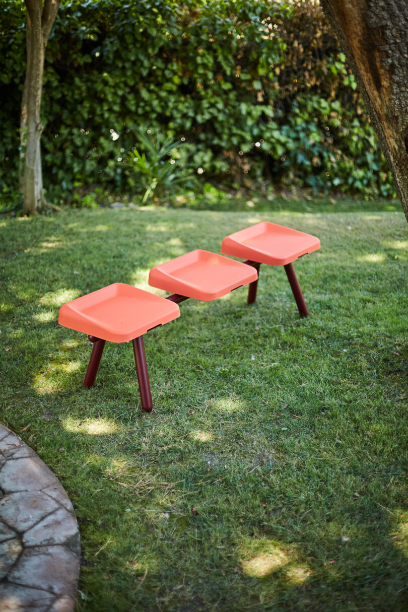 Three orange, square stools with red legs are arranged in a row on a grassy lawn with trees and greenery in the background