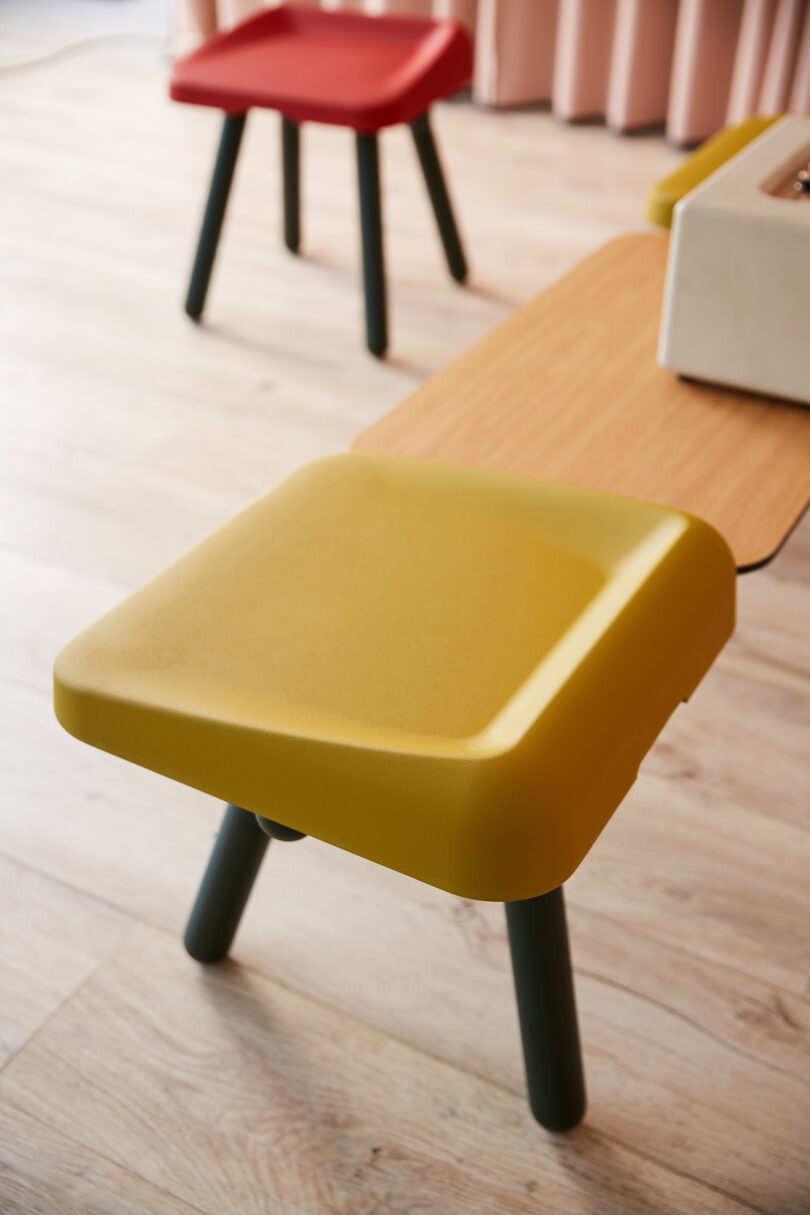 Two stools with green legs and square tops, one yellow and one red, are positioned on a light wood floor next to a wooden table