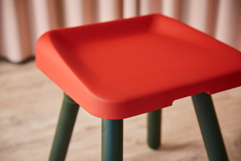 Red square stool with green legs on a wooden floor, in front of beige curtains