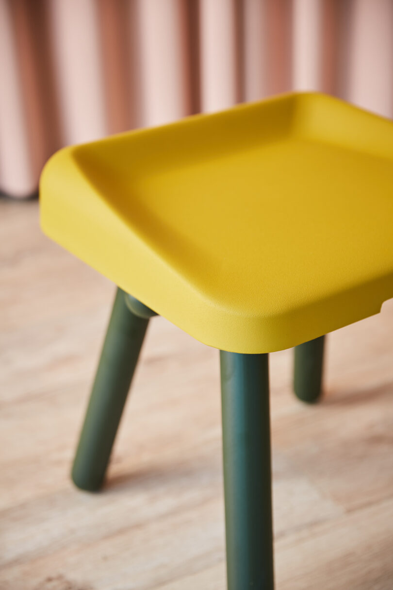 A square yellow stool with green legs on a wooden floor, in front of a blurred pink background