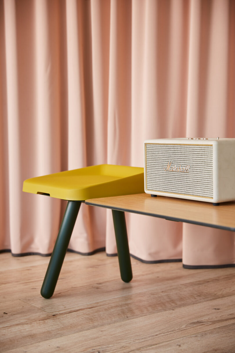 A modern tabletop speaker sits on a wooden table with a mustard yellow tray. A pink curtain serves as the background