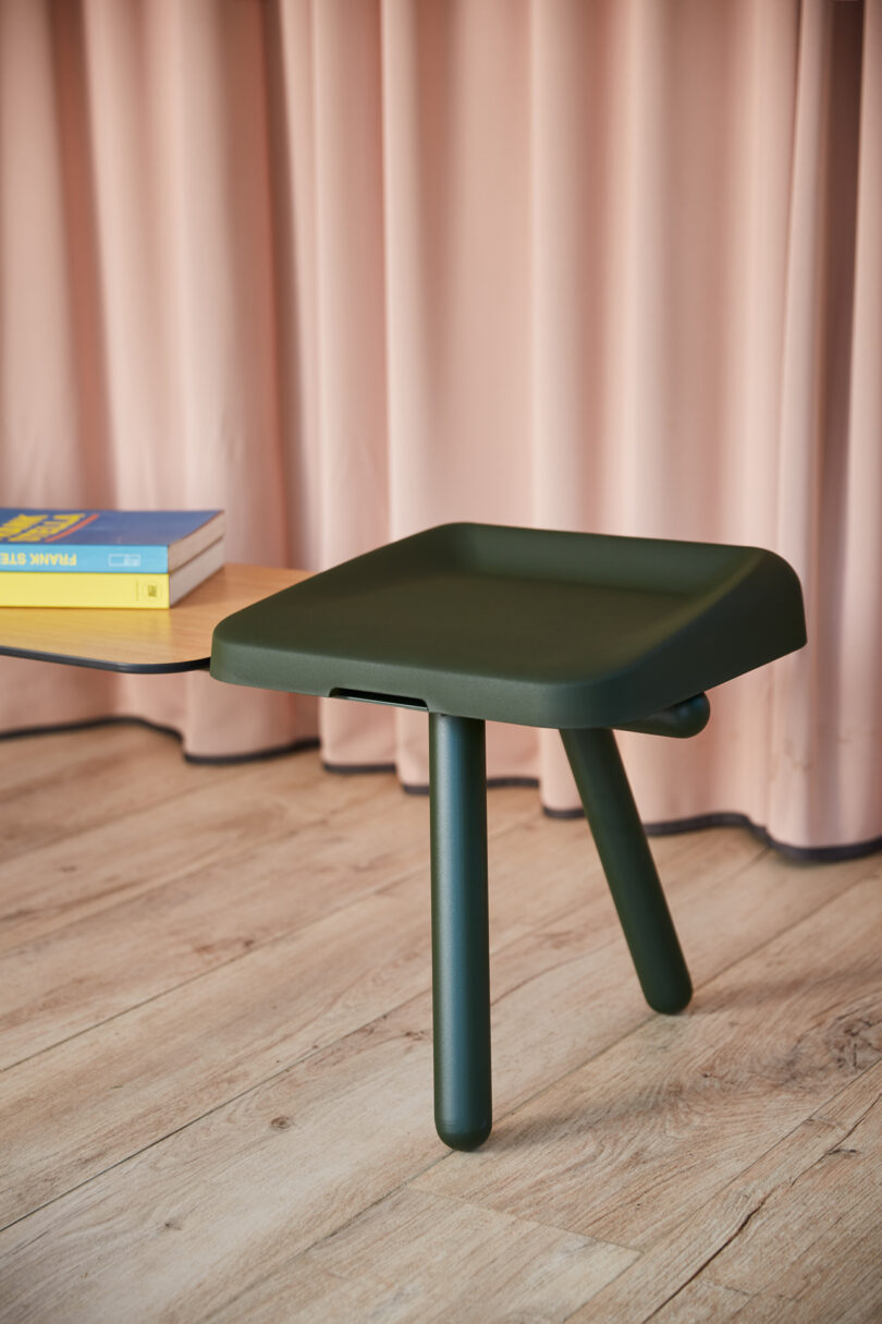 A green three-legged stool sits on a wooden floor in front of pink curtains, with a table holding books partially visible on the left