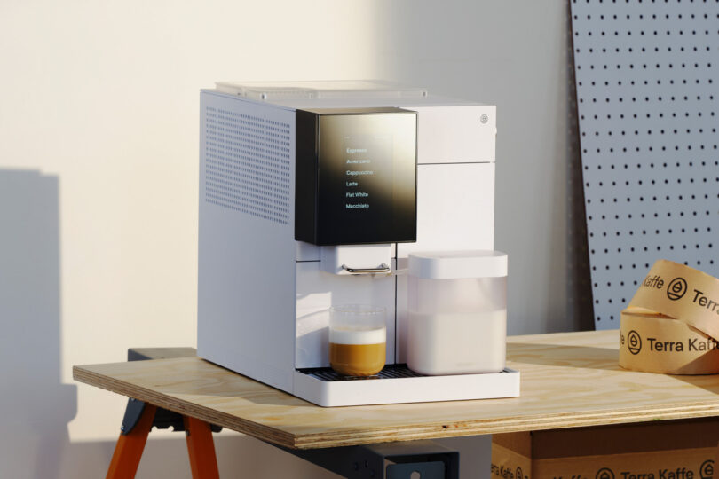 A modern coffee machine on a wooden table brews a cup of coffee, with cardboard boxes in the background.