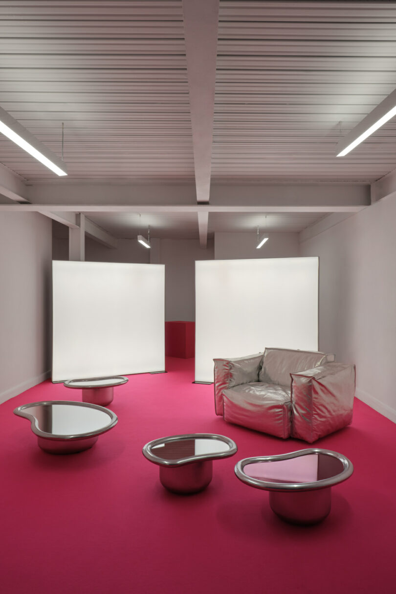 Room with a metallic chair, three reflective tables, and white panels on a pink carpeted floor with overhead lighting