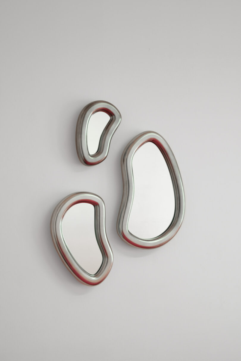 Three irregularly shaped, metallic-framed mirrors are mounted on a white wall, arranged in a cluster