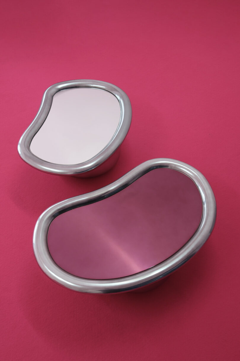 Two kidney-shaped mirrors with metallic frames on a pink background, displaying different shades of reflection