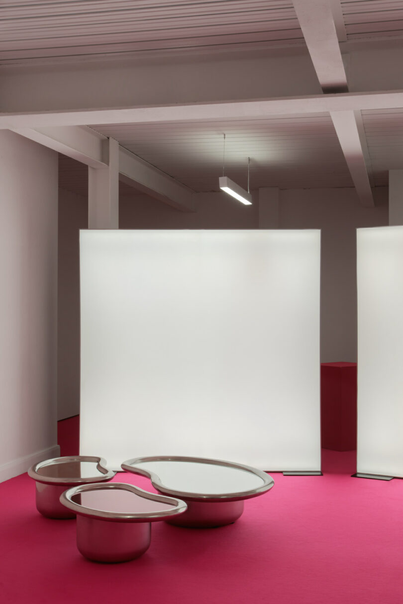 Modern art installation with two reflective, irregular-shaped sculptures on a vibrant pink floor, placed in front of large, illuminated white panels in a minimalist space