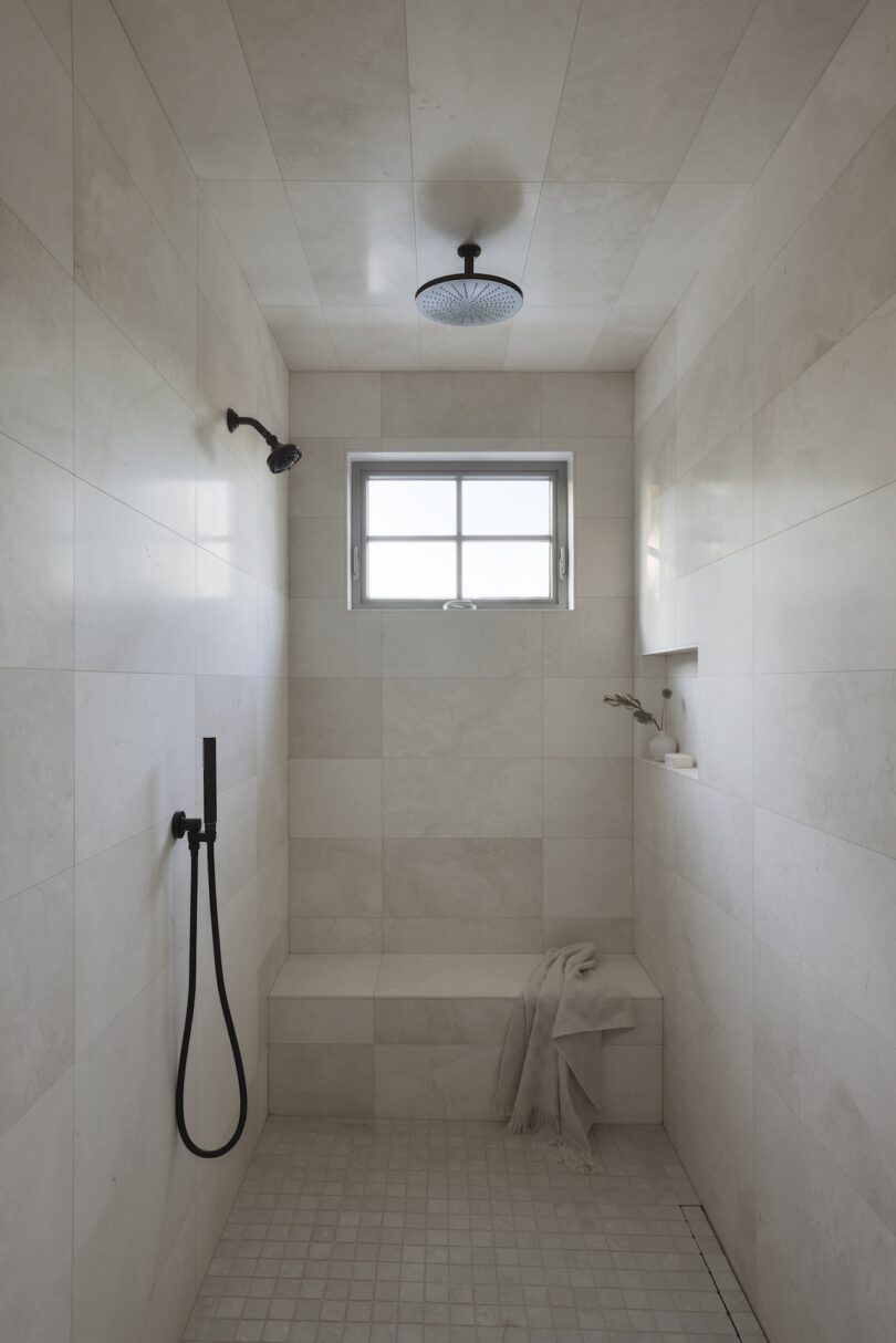 A minimalist shower features beige tiles, a window, a rainfall showerhead, a handheld shower, and built-in bench with a draped towel.