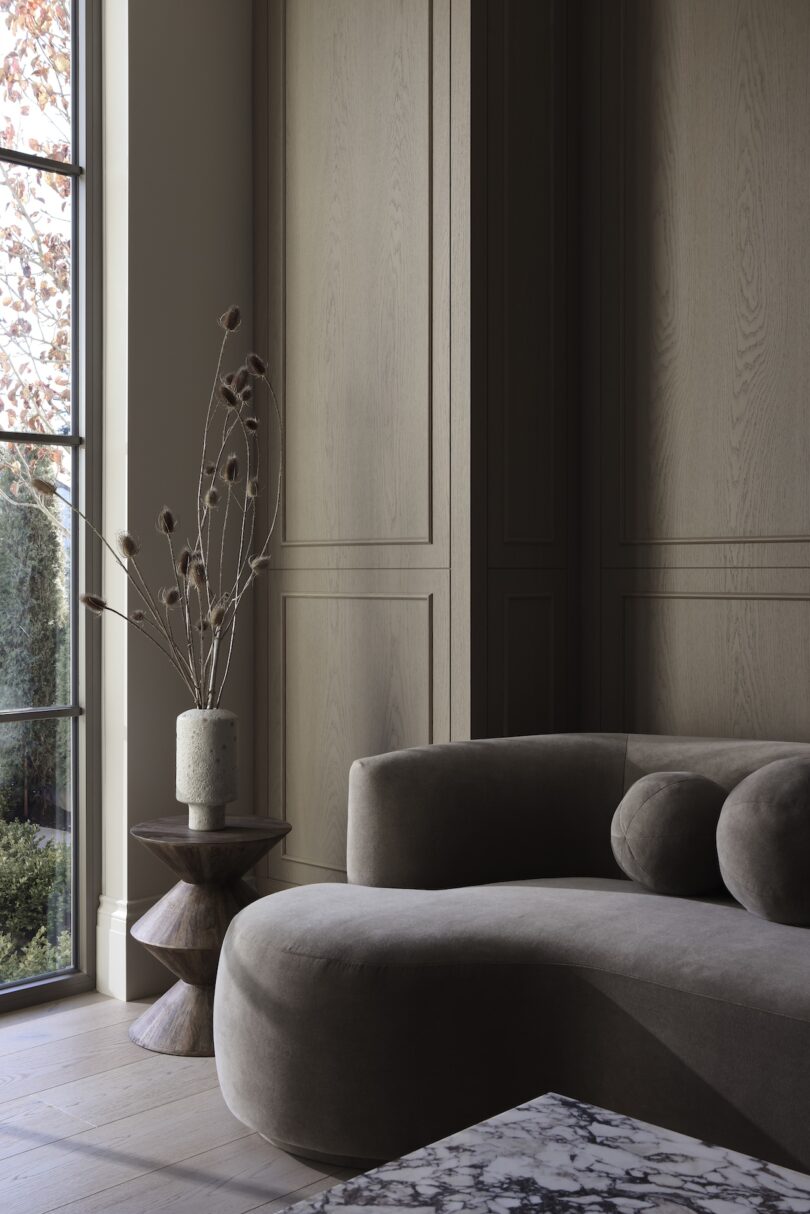 Minimalist living room with a curved gray sofa, a round wooden side table, and a vase with tall dried flowers. Large windows show a glimpse of a garden outside.