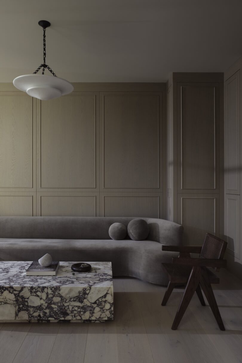 A minimalist living room with a gray sofa, marble coffee table, wooden chair, and a black hanging light fixture against paneled walls.