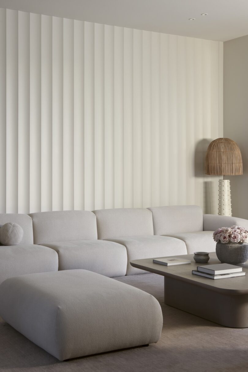 Minimalist living room with a large, beige sectional sofa and ottoman in front of a wavy-textured wall. A woven lamp and a coffee table with books and flowers complete the decor.