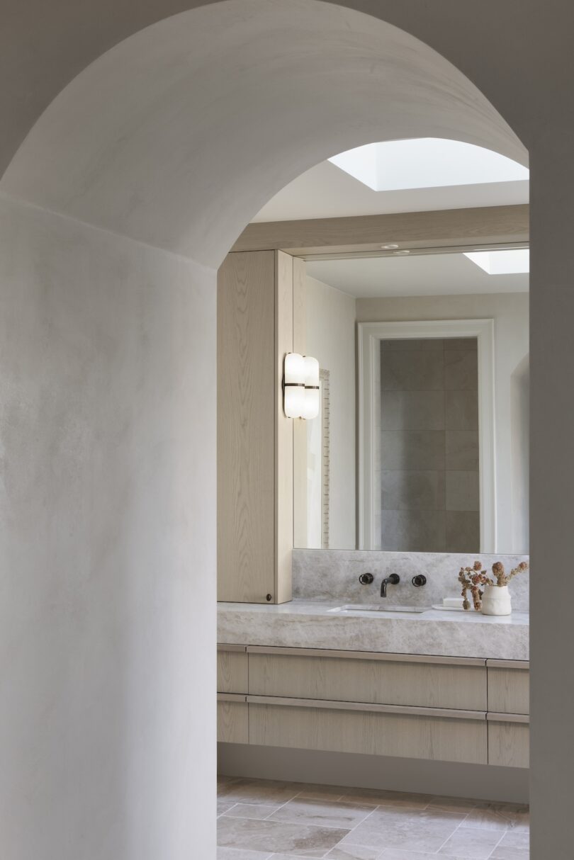 Modern bathroom with arched entrance, featuring a marble countertop, built-in sink, vertical light fixture, and a large mirror. Dried flowers adorn the countertop.