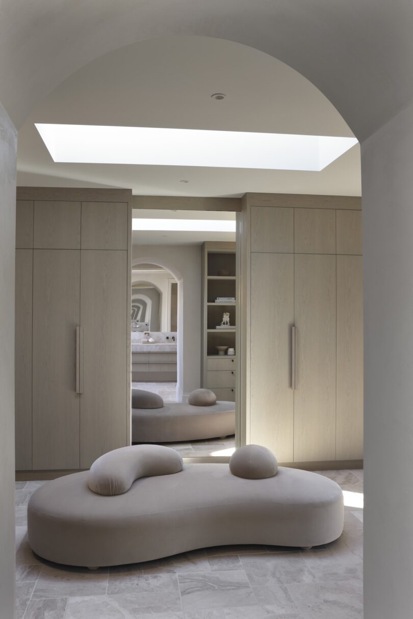 Modern closet with large mirrors, wooden cabinets, and a curved beige bench. Neutral tones and soft lighting create a minimalist atmosphere.