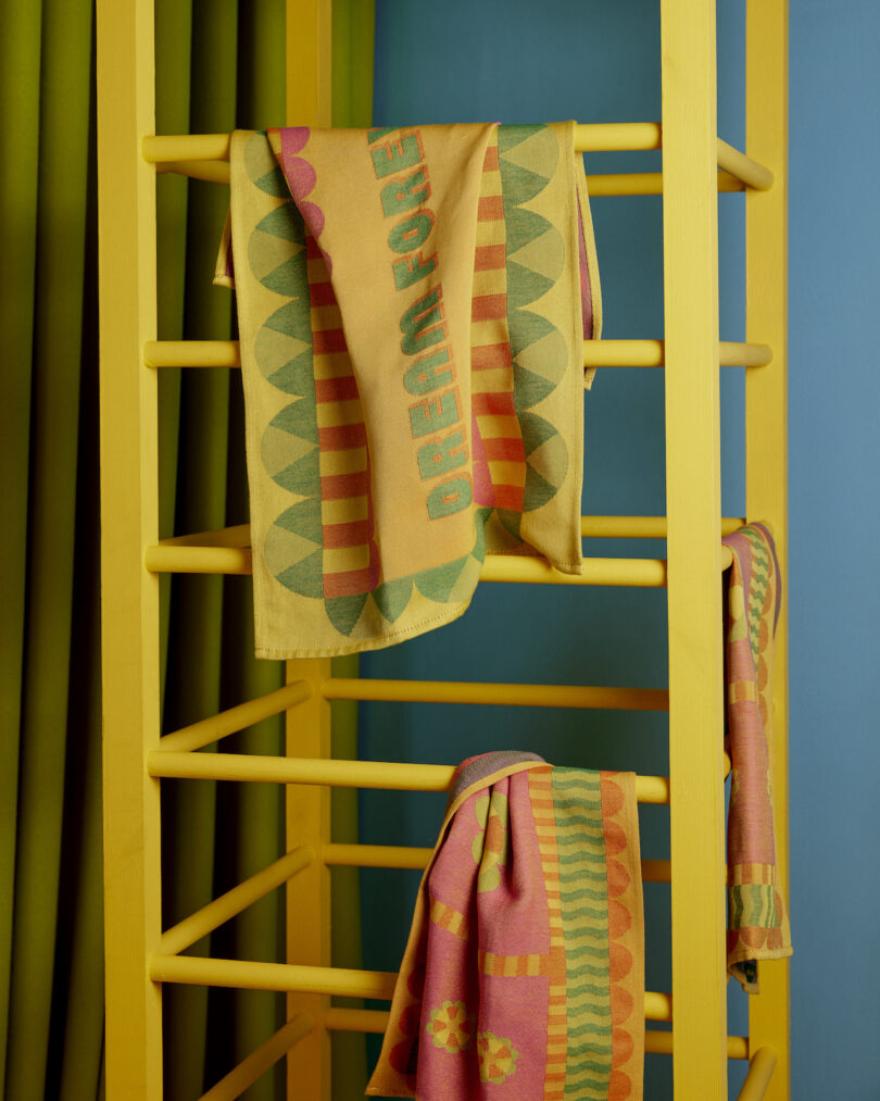 Two towels with geometric patterns hang on a yellow towel rack against a blue wall and green curtain backdrop