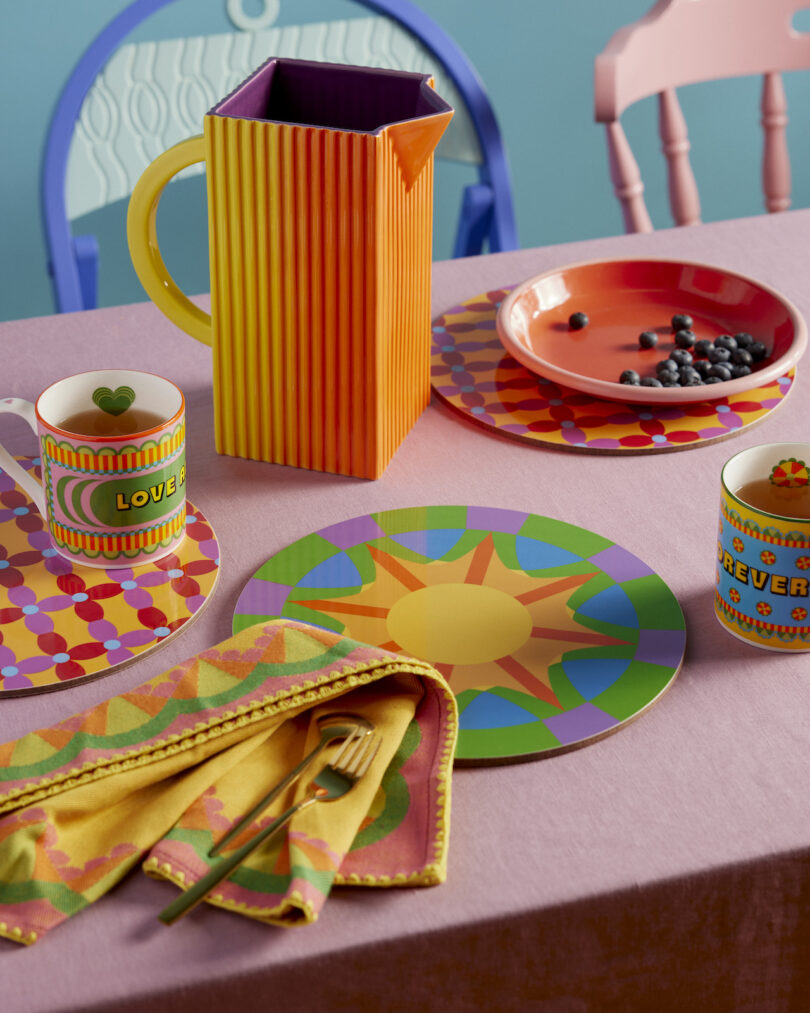 Colorful table setting with patterned plates, mugs, and a striped pitcher on a lavender tablecloth, featuring a few blueberries in a bowl and cutlery on a multicolored napkin