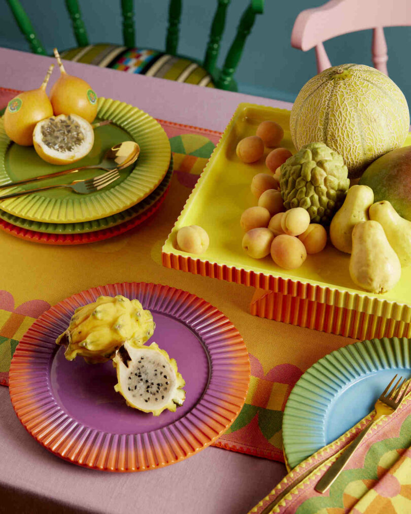 A colorful table setting with vibrant plates, cutlery, and a variety of fruits, including dragon fruit, passion fruit, melon, and pears