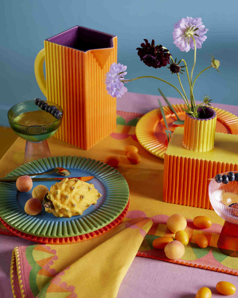 A colorful table setting with vibrant plates, a pitcher, and a vase holding flowers. The table is adorned with fruits, including a horned melon and kumquats, on a patterned cloth