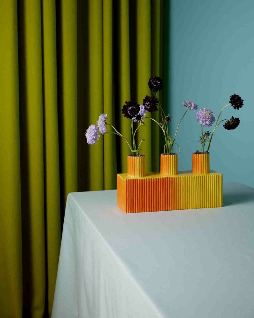 Orange vase with three sections holding purple flowers on a white tablecloth, set against a backdrop of green curtains and a blue wall