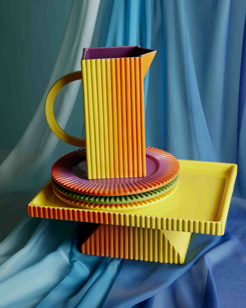 Colorful ceramic tableware, including a tall pitcher, plates, and a tray, with a ribbed texture, arranged on layered blue cloth