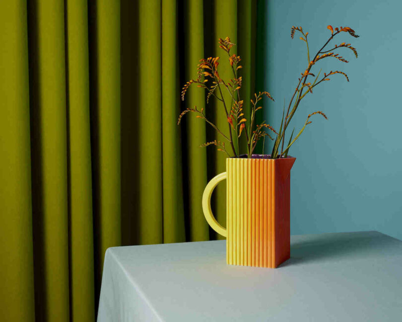 A colorful striped pitcher with sprigs of orange flowers sits on a white table against a backdrop of green curtains and a teal wall