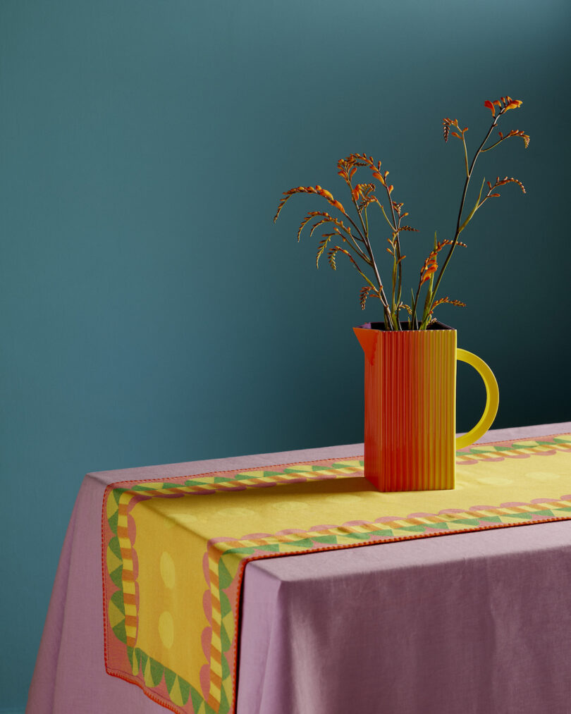 A colorful pitcher with flowers sits on a table covered by a pink cloth and yellow runner against a teal background