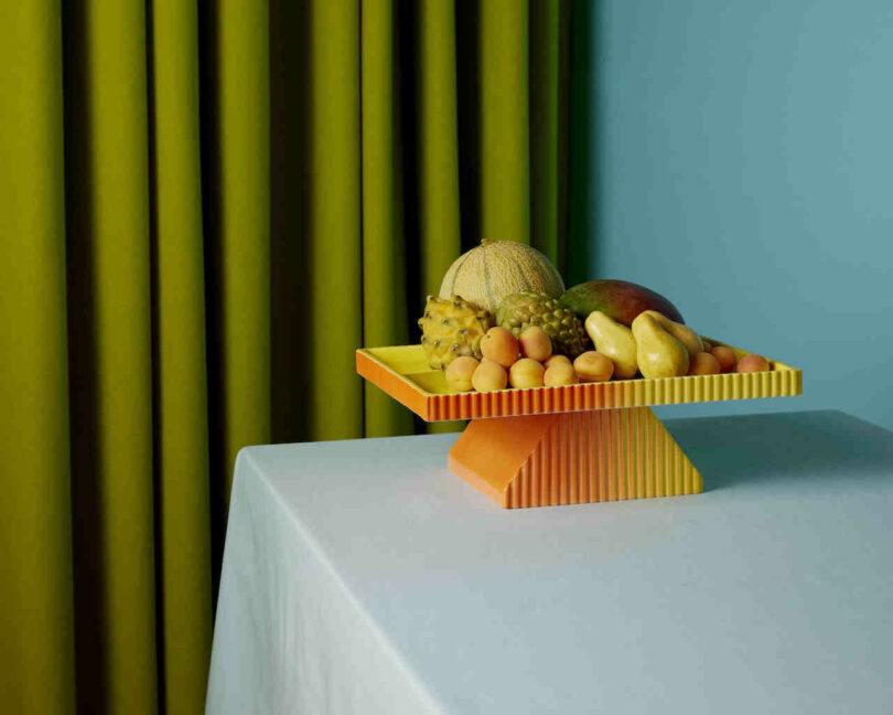 A geometric fruit stand with a variety of fruits, including a melon, mango, and small yellow fruits, is placed on a white tablecloth. A green curtain and blue wall are in the background