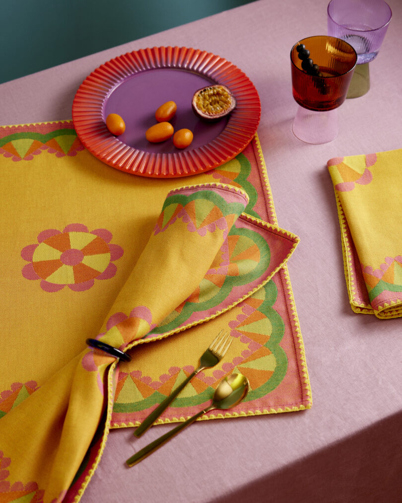 A vibrant table setting with colorful textiles, a purple plate with kumquats and half a passion fruit, gold cutlery, and gradient glasses on a pink tablecloth