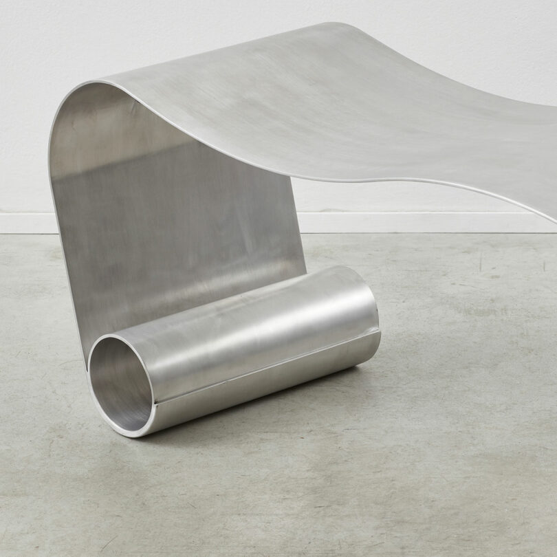 A modern, minimalist stainless steel bench with a smooth, curved design sits on the gray concrete floor, embodying an aesthetic reminiscent of an Alu Archive for Space.