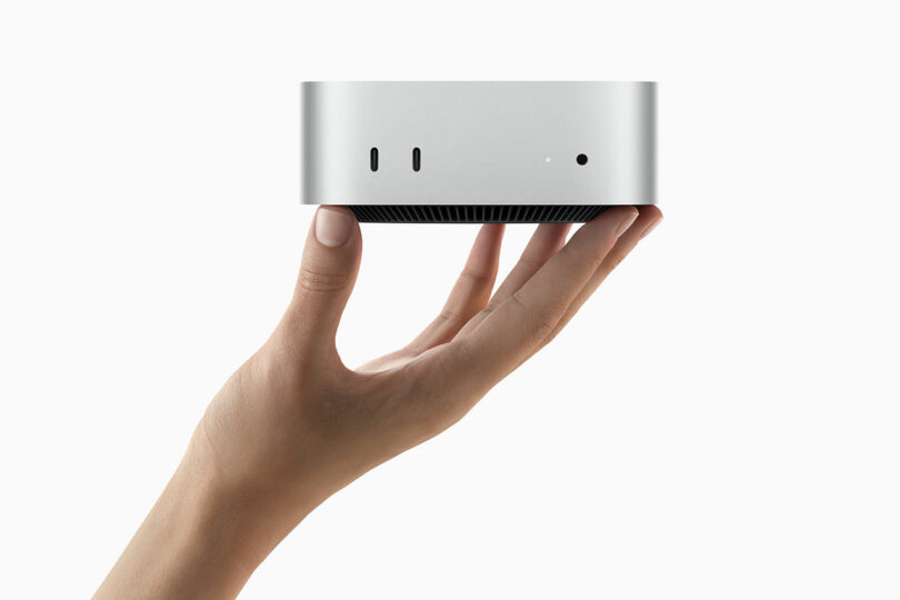 A hand holds a compact silver Mac Mini device with ports on the front, set against a plain white background.