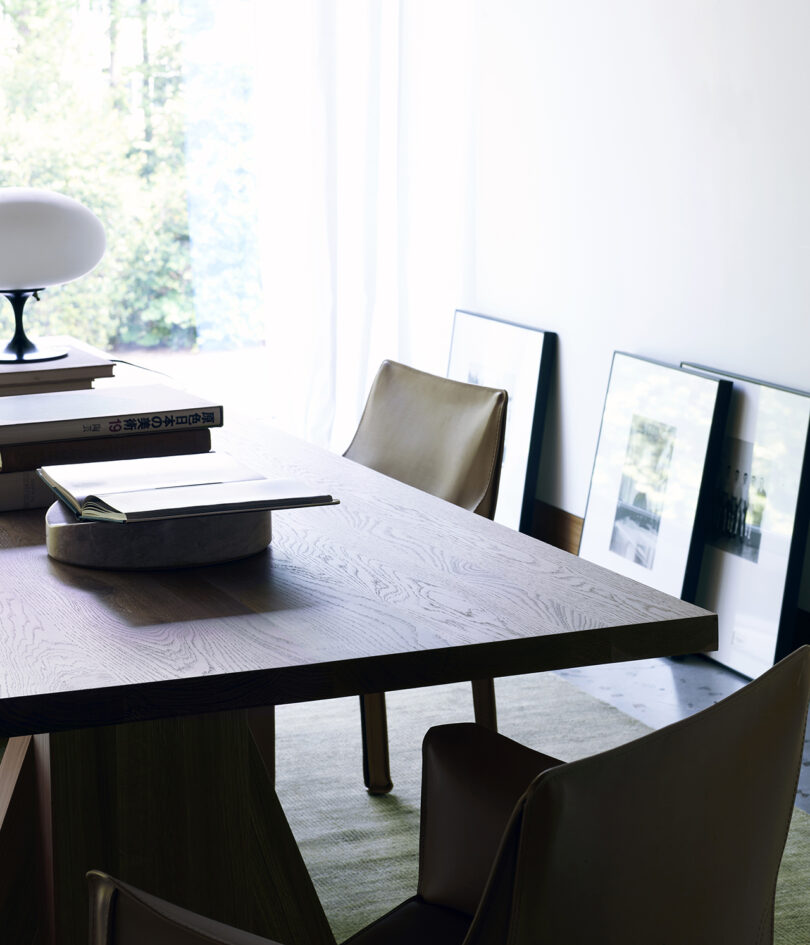 A wooden dining table with chairs, topped with books and a decorative object. Framed artwork is leaning against the wall in a light-filled room.