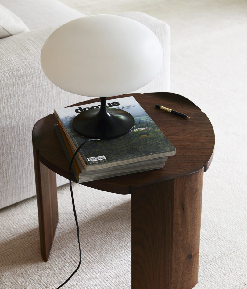 A white lamp sits on a stack of magazines on a dark wooden side table next to a beige sofa. A pen is placed on the table.