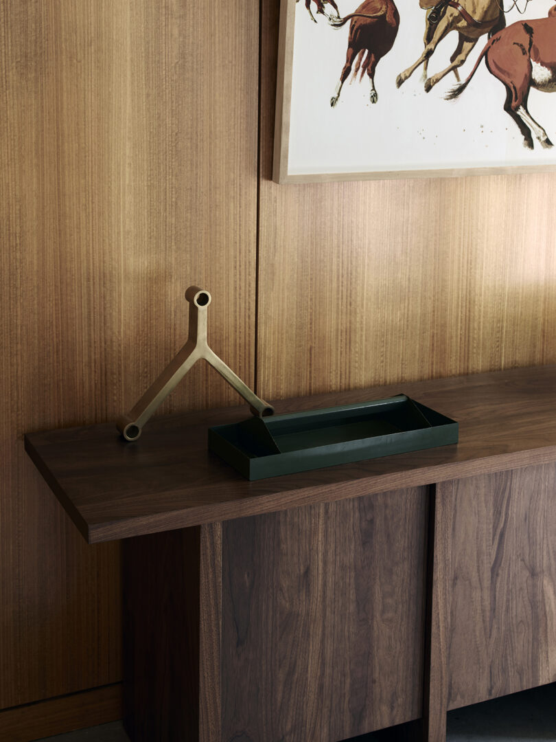 A wooden cabinet with a green tray and a geometric metal sculpture in front of a wood-paneled wall and a painting of horses.