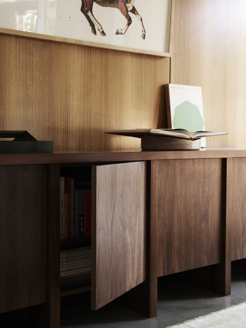 A wooden cabinet with books, a partially open door, and an open book on top. An artwork featuring a horse is on the wall above.