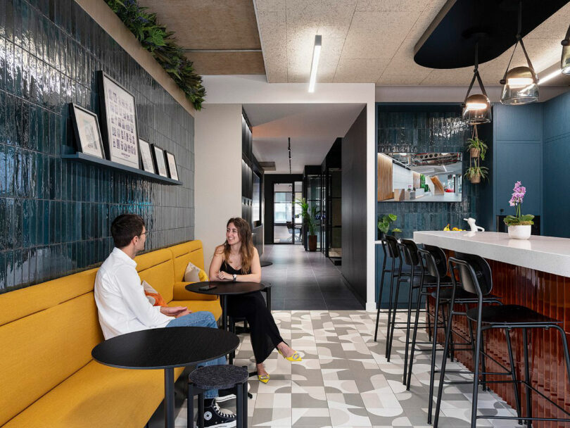 A man and woman sit at a small table in a modern, stylish office break area with green tiles, a long counter, and barstools.