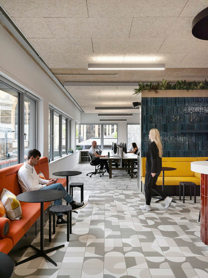 Modern open office space with people working and relaxing. Includes seating area, workstations, large windows, and geometric patterned flooring.