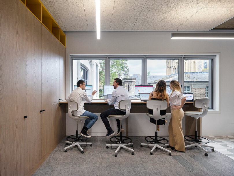 Four people sit on high stools at a desk by a window, working on laptops in a modern office space.