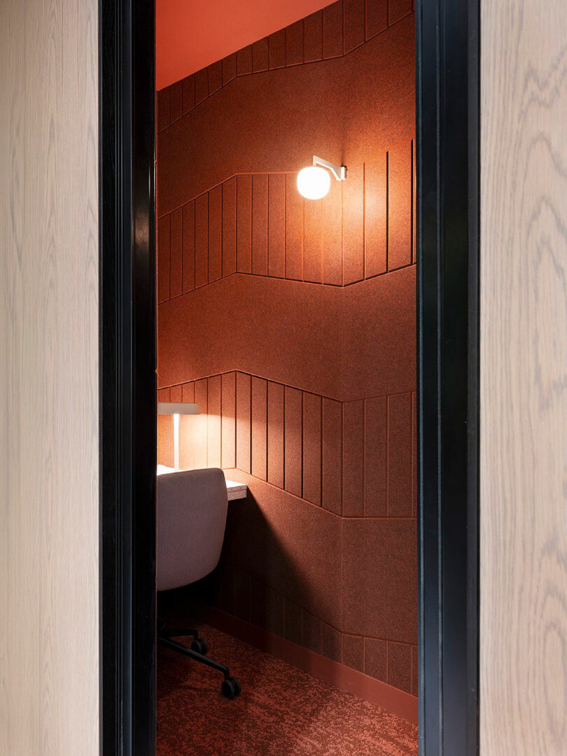 A small, modern study nook with red walls, a chair, desk, and wall-mounted lamp.