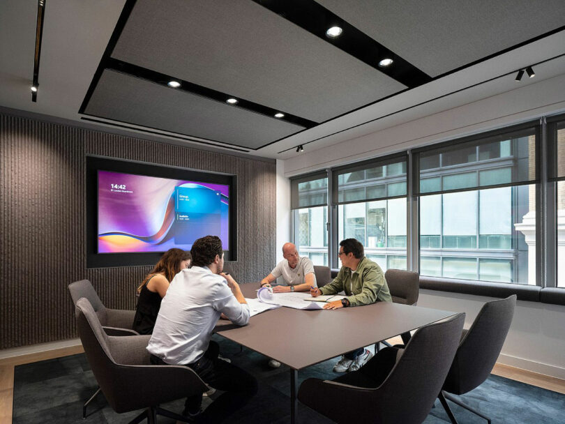 Four people at a conference table reviewing documents in a modern meeting room with a screen displaying colorful graphics.