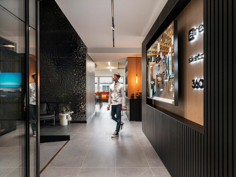 A person walks through a modern office hallway with gray tiles and a large digital display on the wall.