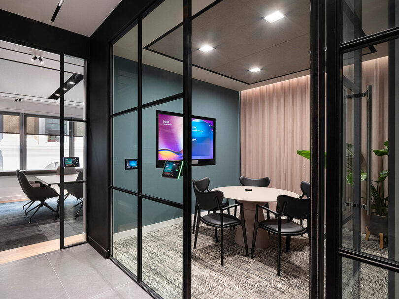 Modern meeting room with glass walls, a round table, four chairs, and digital screens on walls.